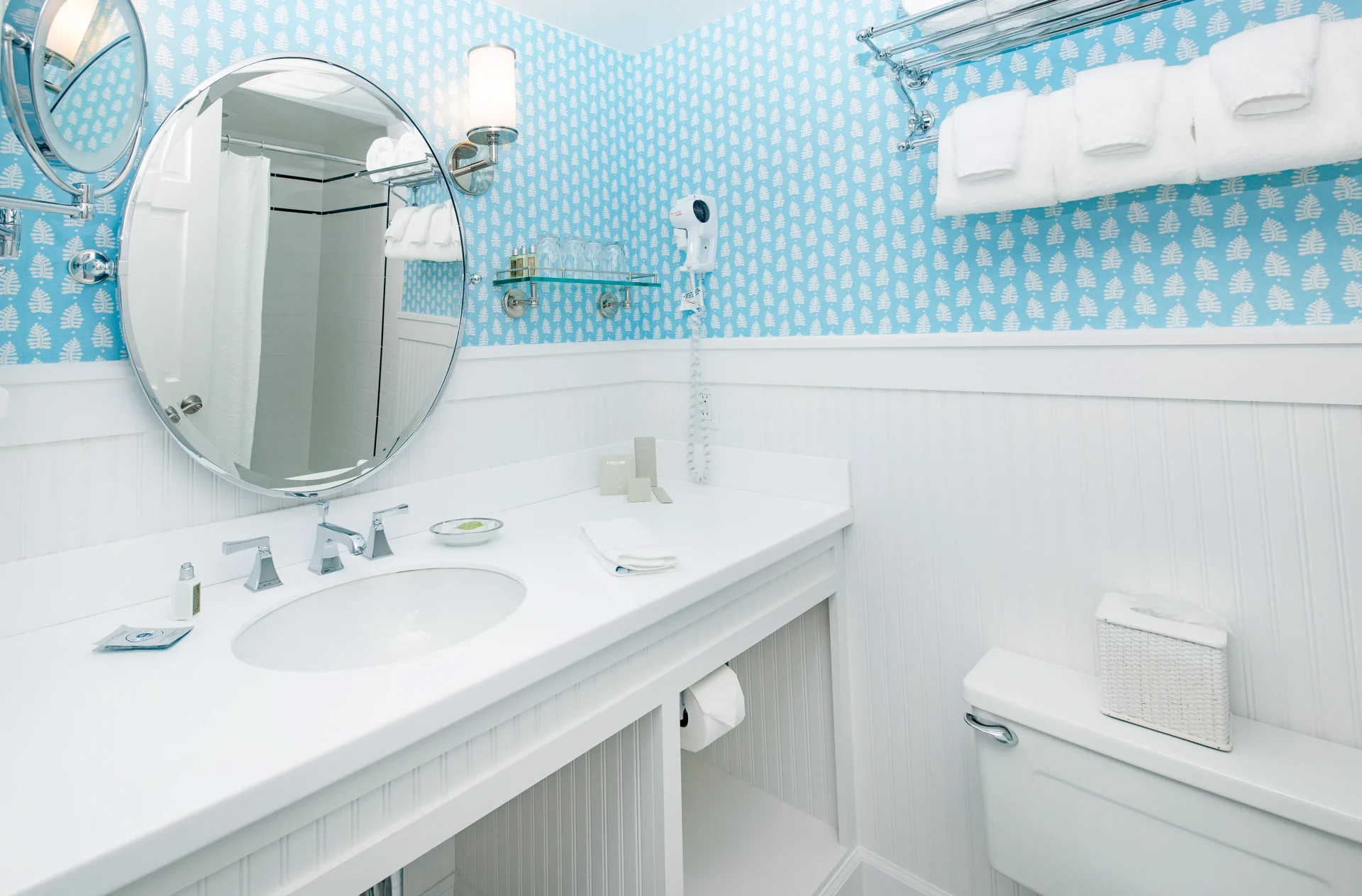 A white bathroom with blue wallpaper with the view of the sink, mirror and toilet.