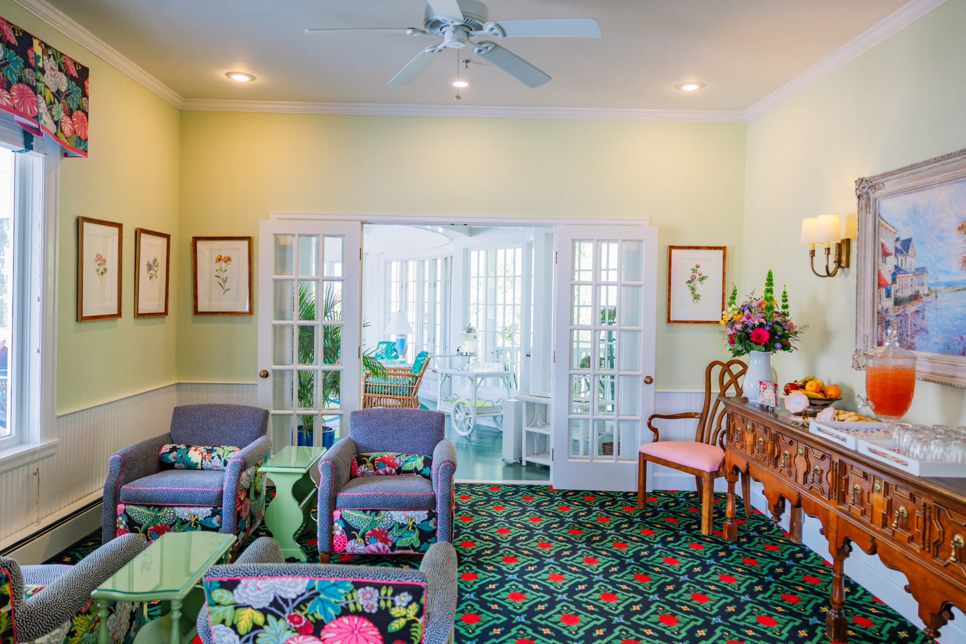 A green, black and red patterned carpeted room with purple chairs, wood table with available punch looking out onto many window walkways.