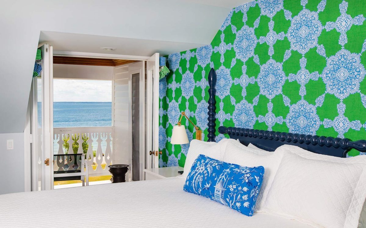 A white bed in a blue and green patterned room looking at a balcony that looks out onto Lake Michigan.