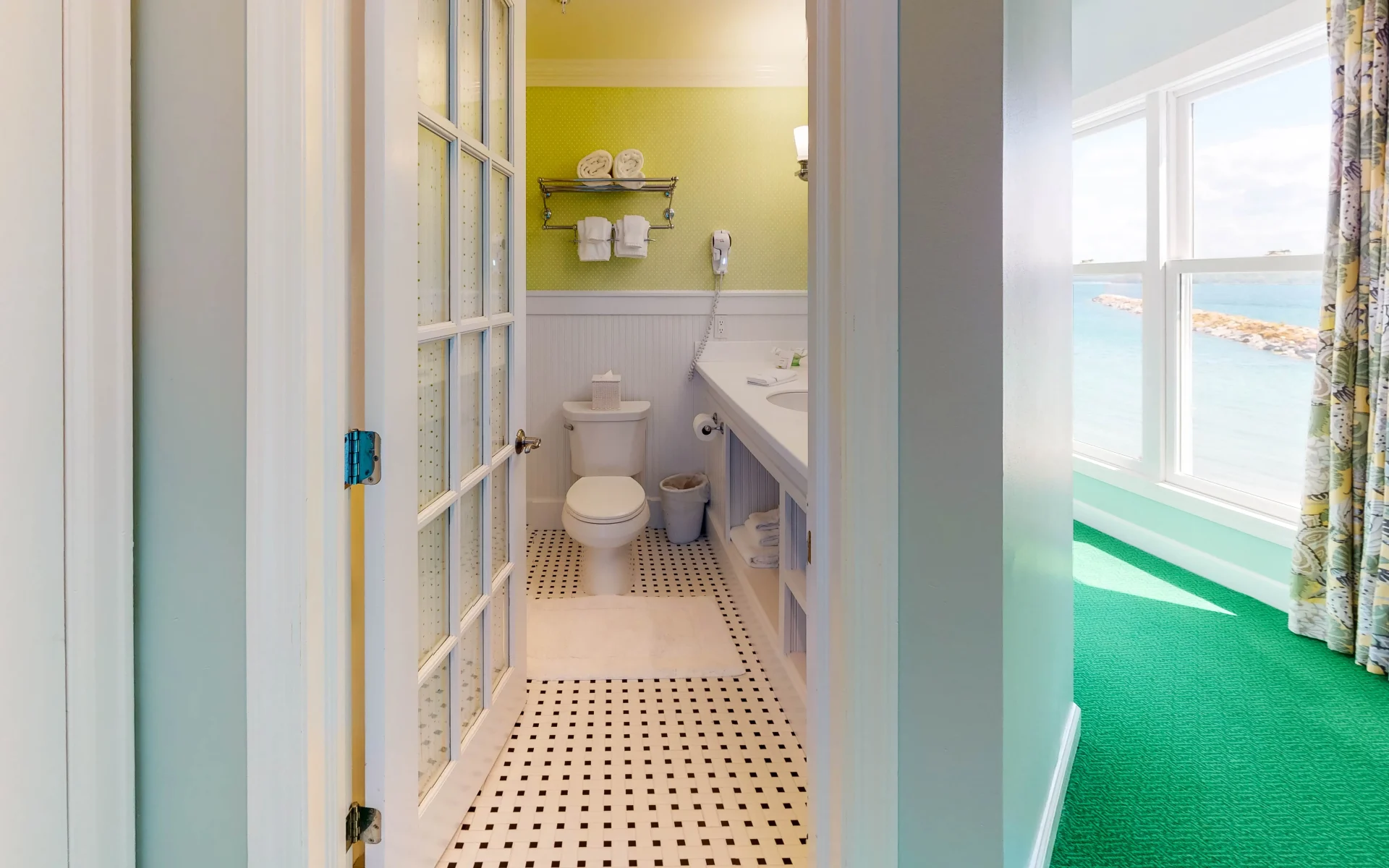A green and white bathroom with white and black tiled floor with view of the toilet and entry door.