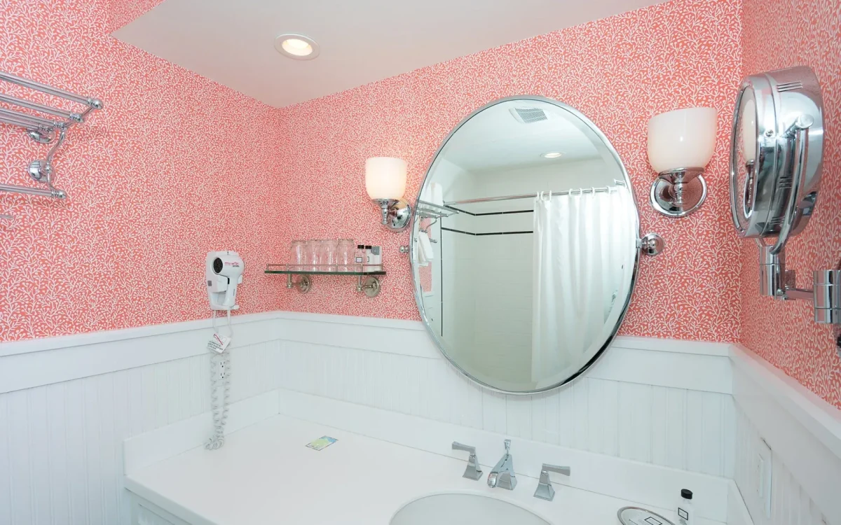 A white bathroom with pink accent wallpaper looking onto the sink and mirror.