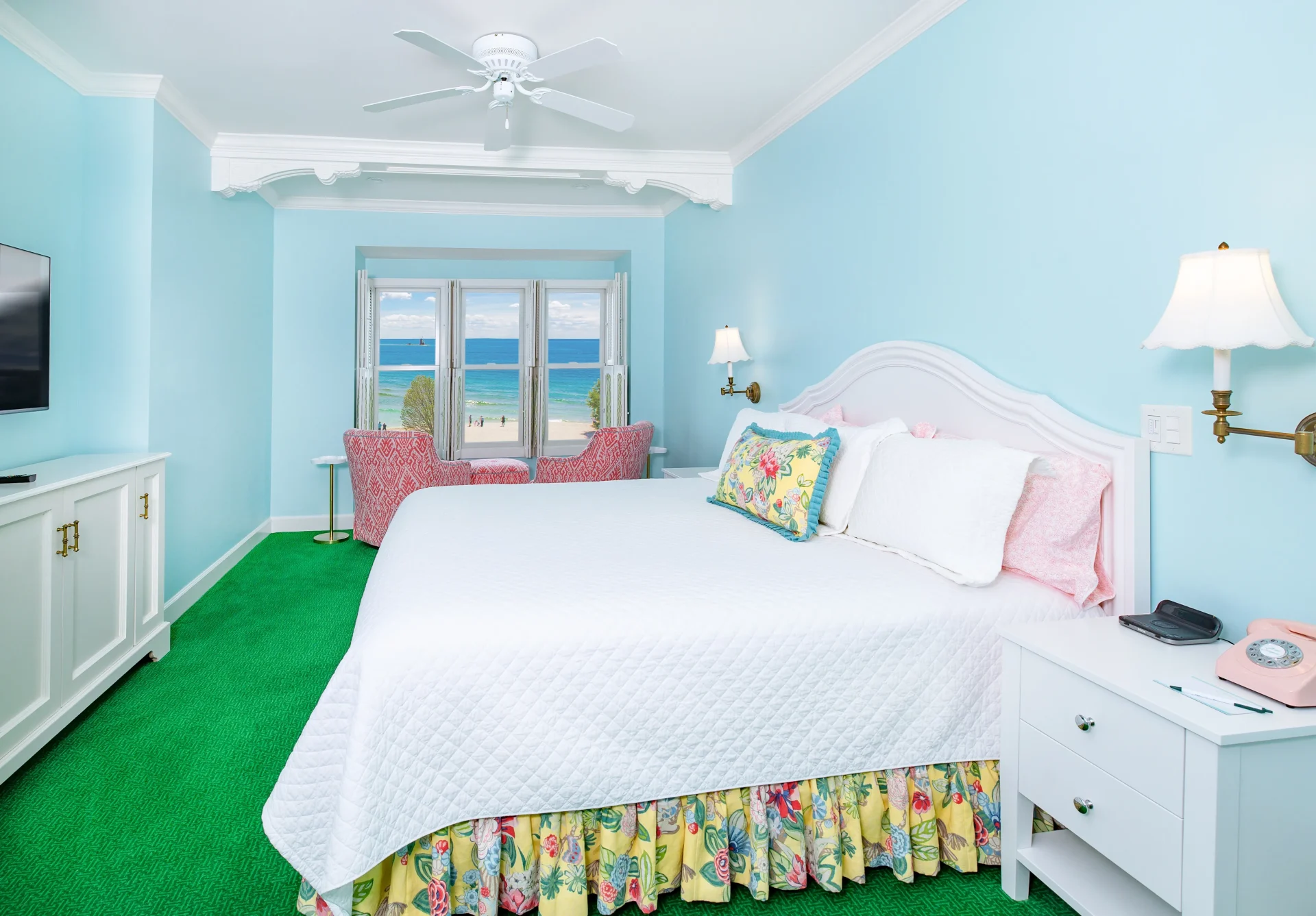 White bed in a blue painted room with green carpet, ceiling fan, and windows looking out onto a beach.