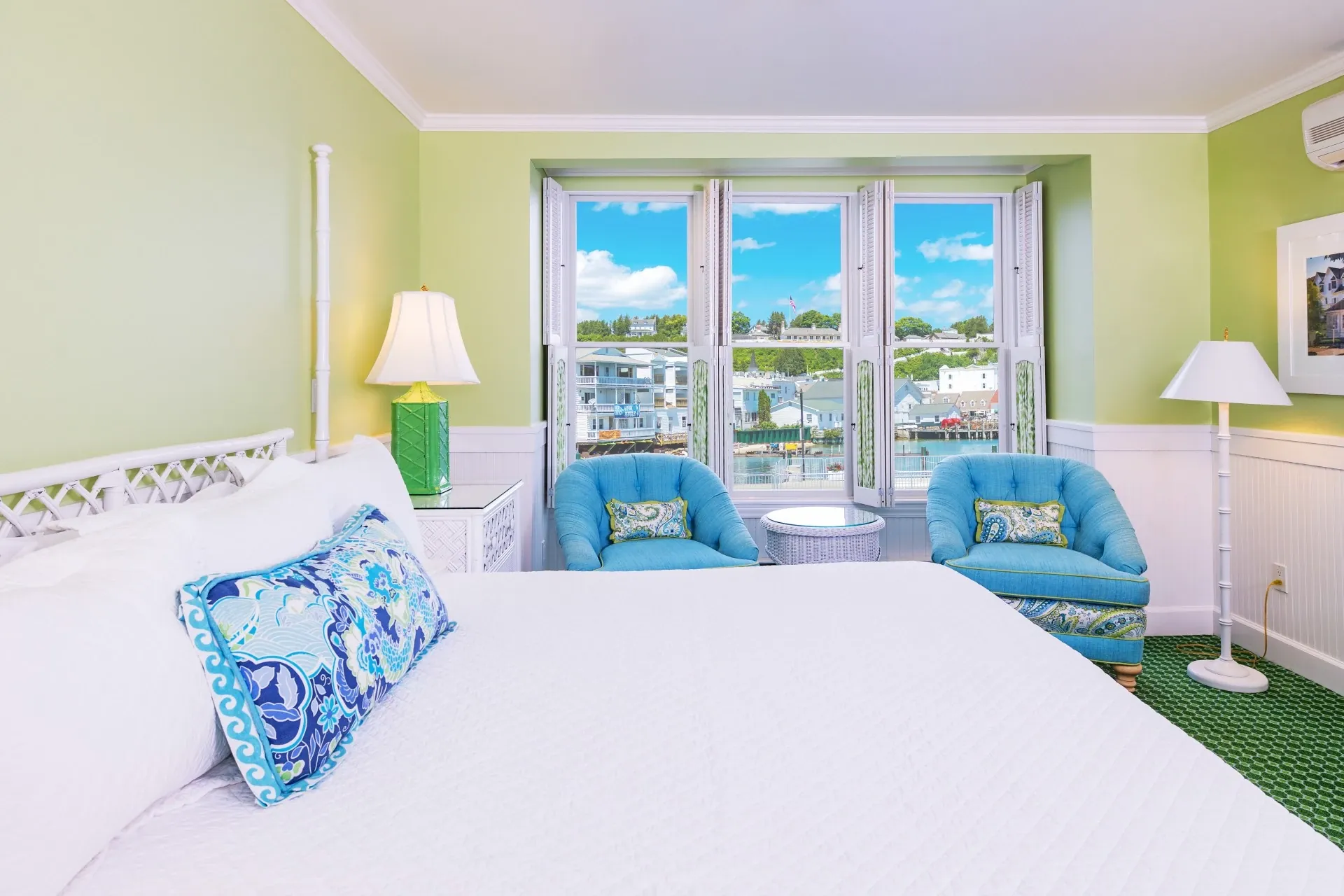 A white bed and a green room with a lamp, windows, and chairs