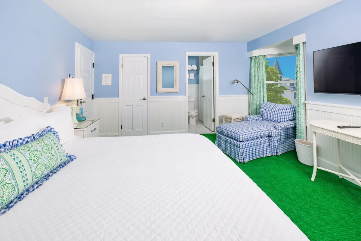 A white bed and a blue room with a blue chair with windows and TV.