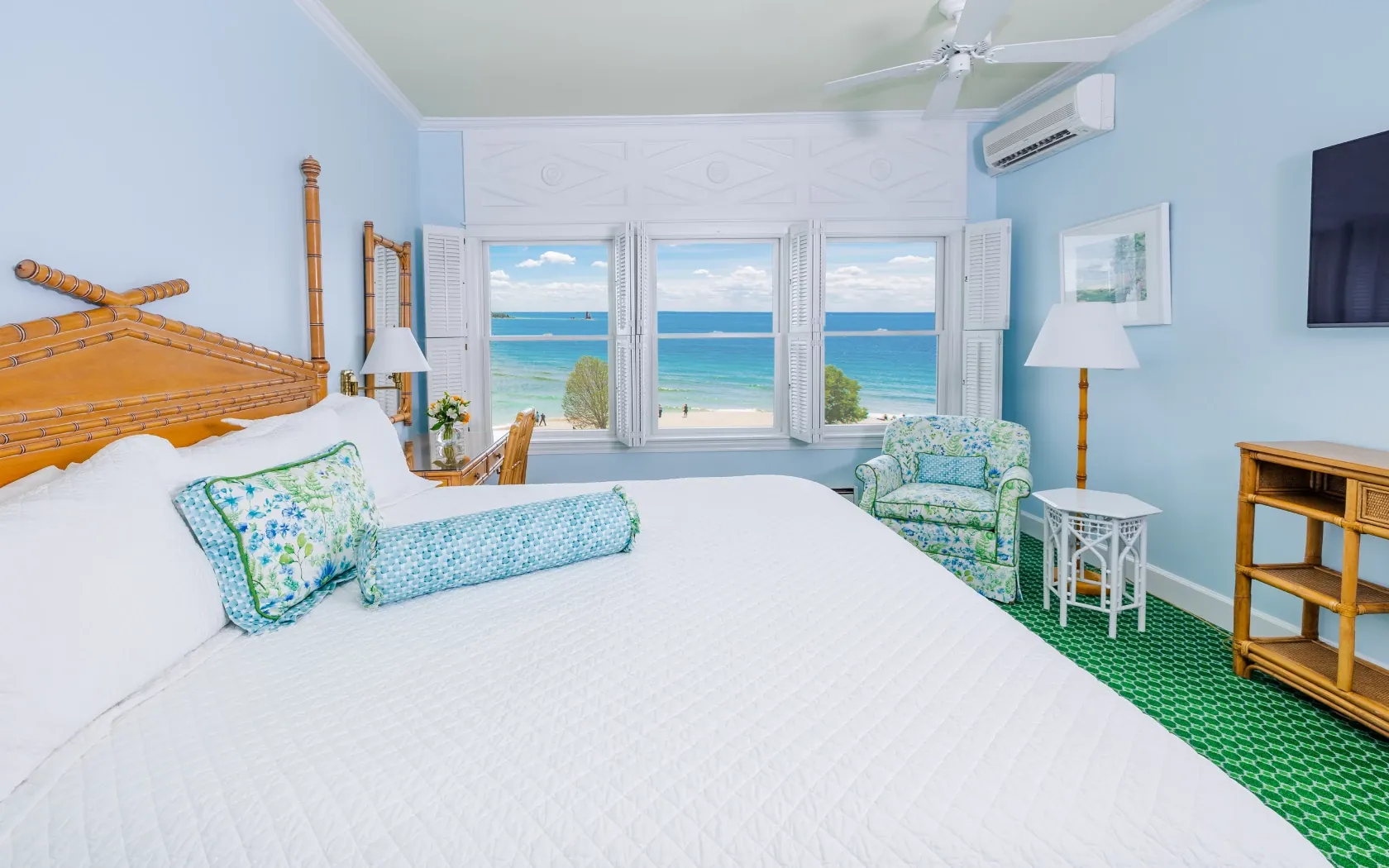 A white bed and a blue room with a ceiling fan, with green pattern carpet, a pink chair, and three windows looking out onto a beach