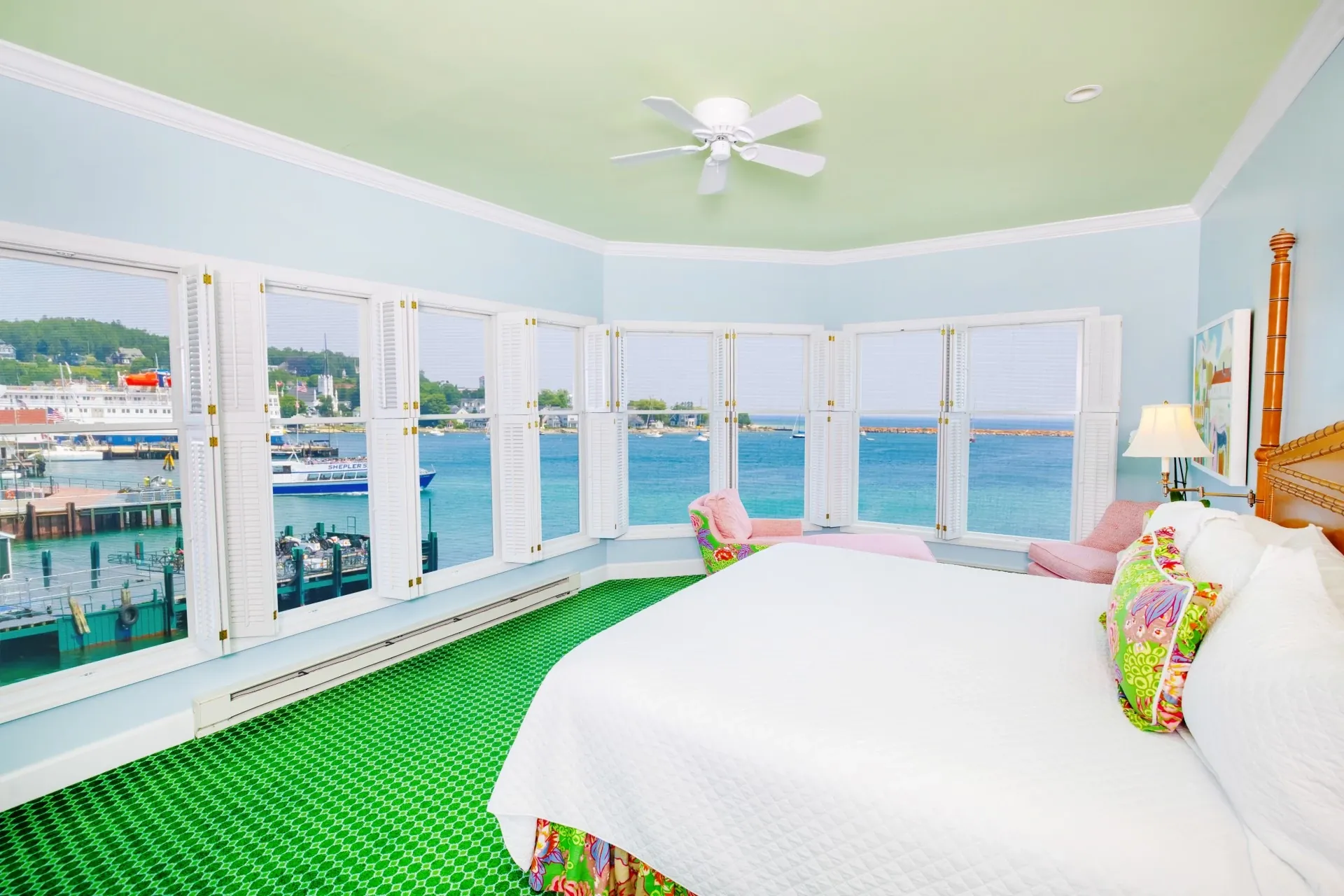 A white bed in a blue room with green carpet with a ceiling fan and many windows looking out onto the Lake.