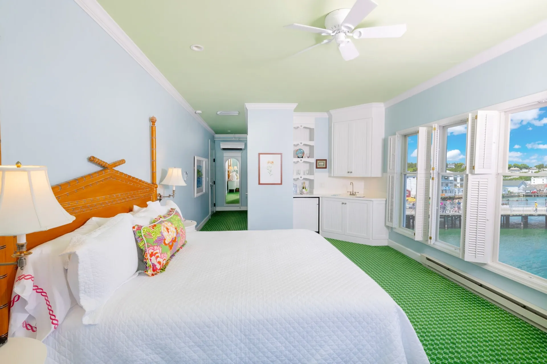 White bed in a blue room with green carpet with kitchenette with view of ceiling fan and windows out onto a lake.