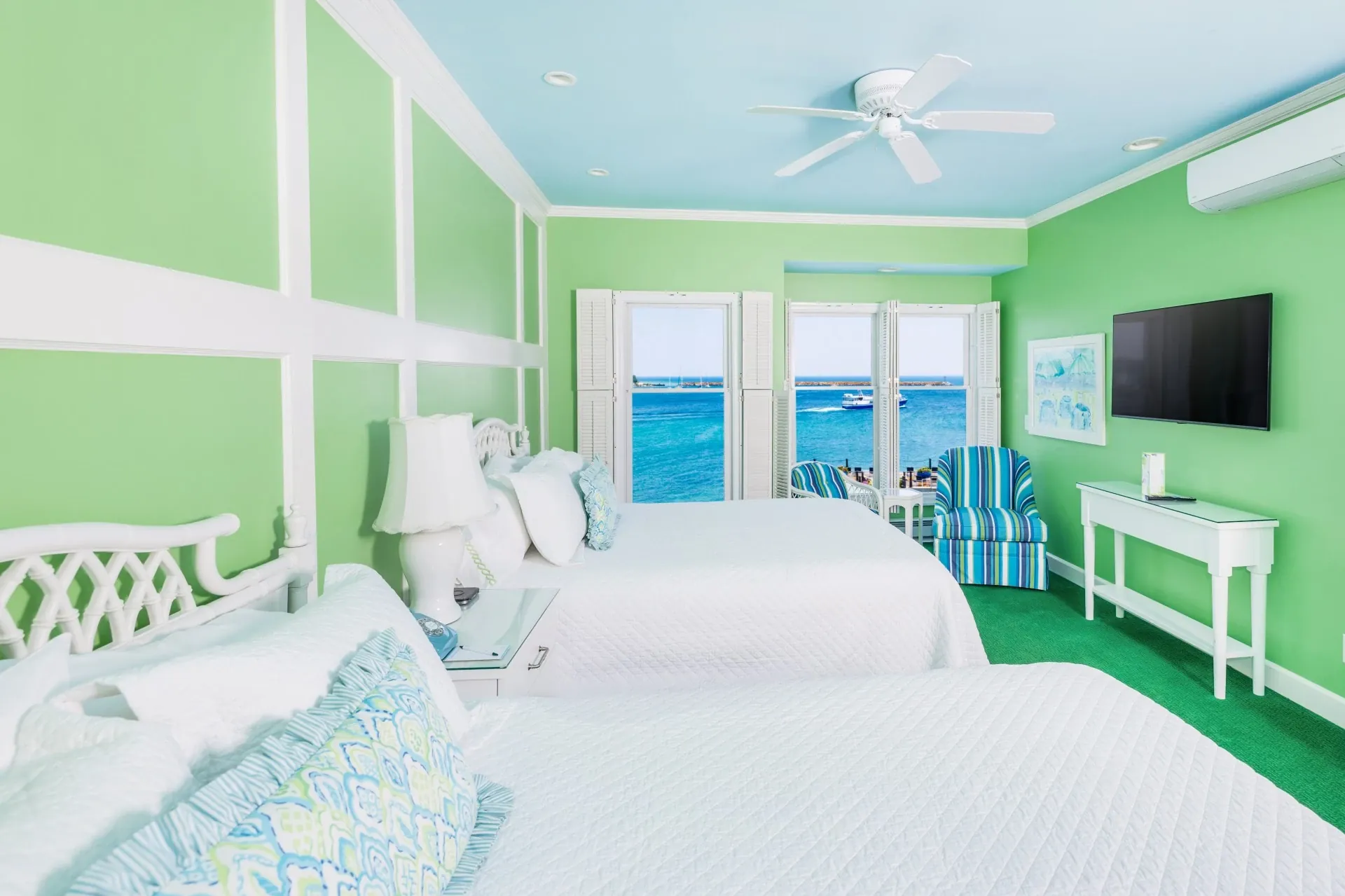 Two queen beds in a green room with ceiling fan with blue accent ceiling with three windows looking out onto the lake.