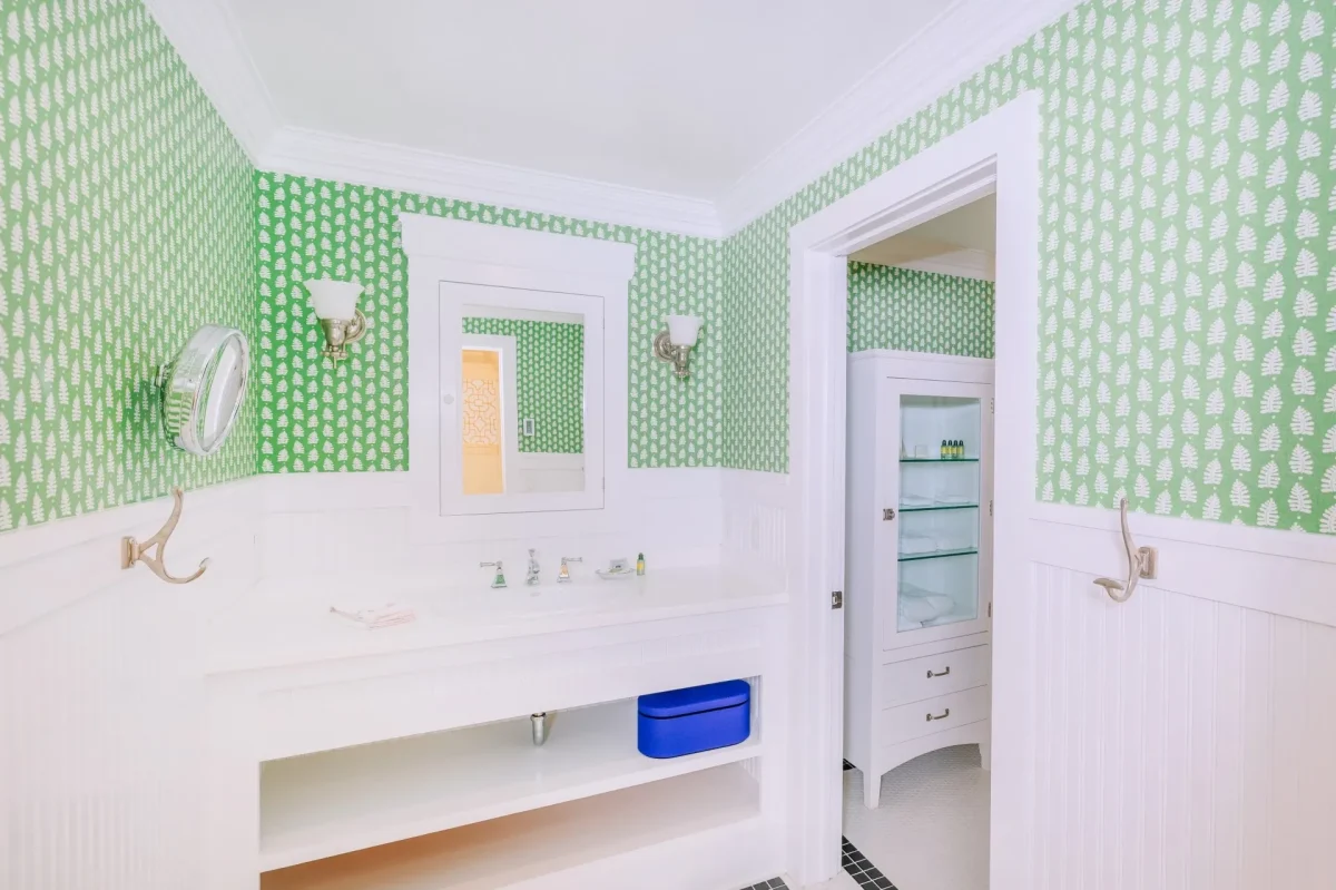 A white bathroom with green patterned wallpaper with view of the sink, mirror, and entryway.