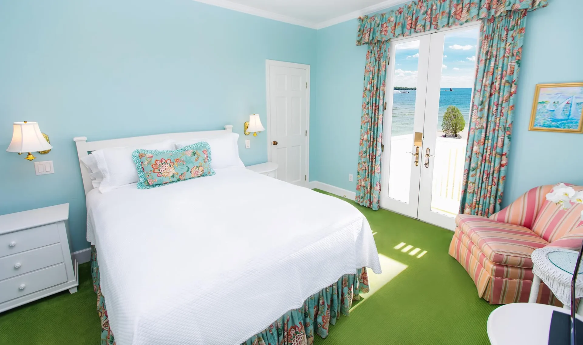 A white bed in a blue room with green carpet, with curtain, with Balcony overlooking a beach.